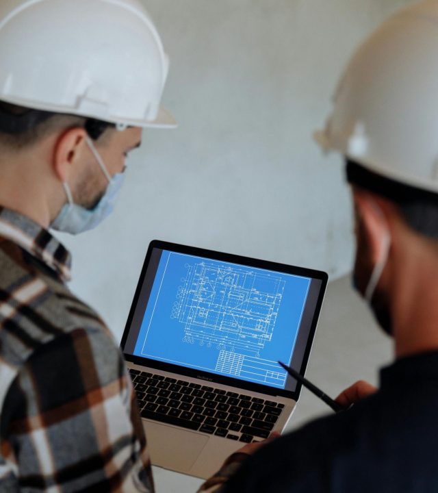 Engineers wearing hard hats and masks review a construction blueprint on a laptop indoors.